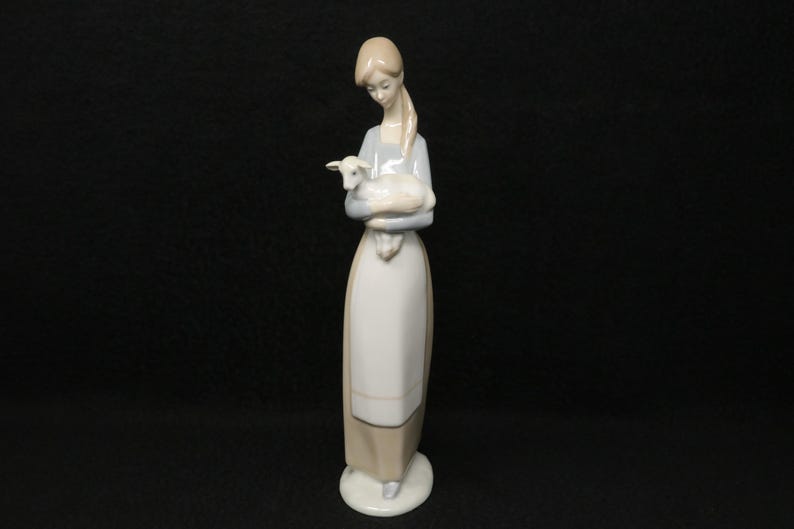 May include: A porcelain figurine of a woman holding a lamb. The woman is wearing a long, light-colored dress with a matching apron. The lamb is white with a light blue hue. The figurine is on a white circular base.