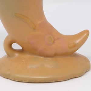 May include: A ceramic vase shaped like a crescent moon with a pink flower design. The vase is a light brown color and has a small hole on the side.