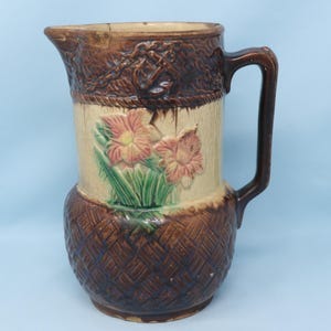 May include: Antique ceramic pitcher with a brown handle and spout. The pitcher features a floral design with pink and yellow flowers on a cream-colored background. The base has a woven basket pattern.