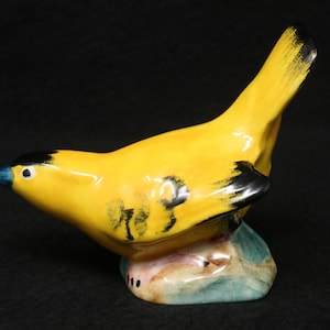 May include: A yellow ceramic bird figurine with black accents and a blue beak, perched on a green and brown base.