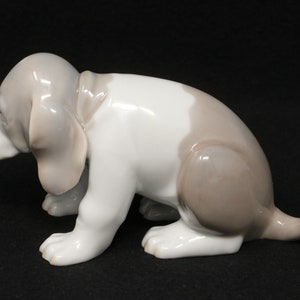 May include: A porcelain figurine of a brown and white puppy sitting on its hind legs with its head down. The puppy has floppy ears and a long tail.