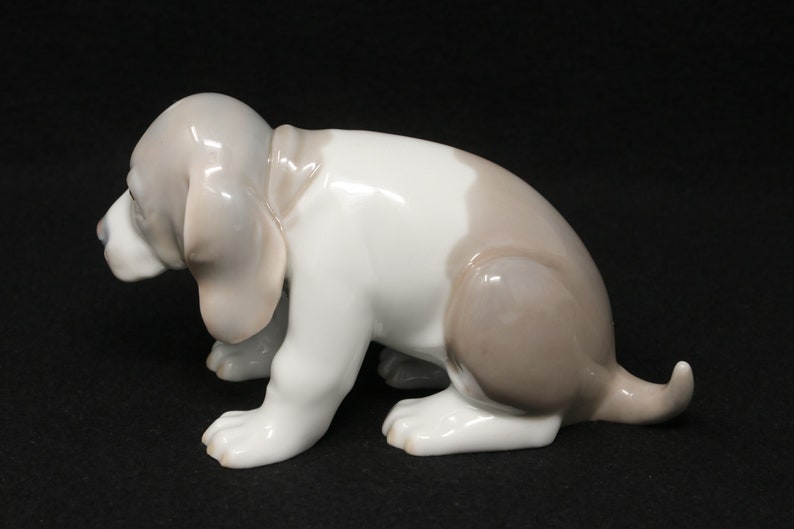 May include: A porcelain figurine of a brown and white puppy sitting on its hind legs with its head down. The puppy has floppy ears and a long tail.