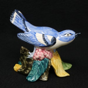 May include: A blue and white ceramic bird figurine perched on a green and yellow floral base. The bird has a black eye and a long, pointed beak.