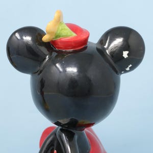 Puede incluir: Una figura de Mickey Mouse negro con un sombrero rojo y un acento amarillo y verde. La figura tiene grandes orejas negras y un lazo rojo. La figura es brillante y parece ser de cer&aacute;mica.