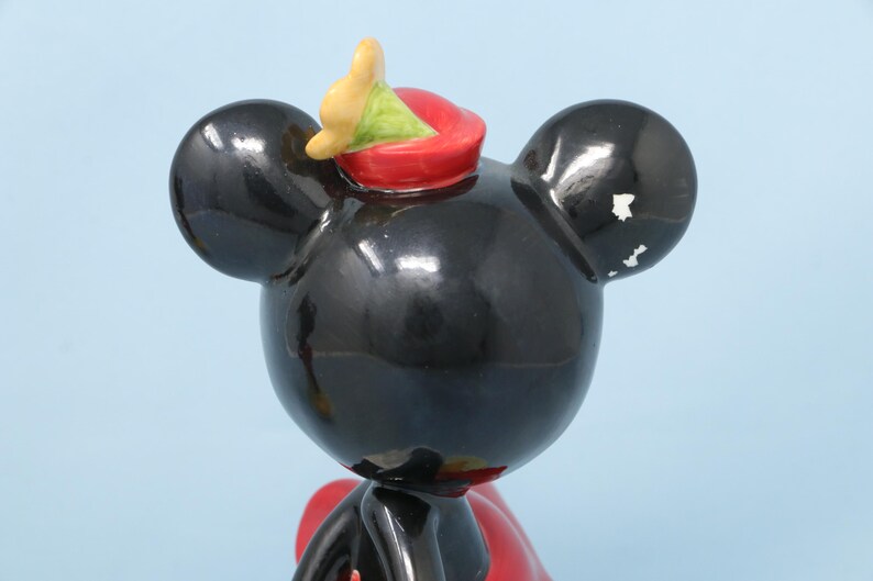 Puede incluir: Una figura de Mickey Mouse negro con un sombrero rojo y un acento amarillo y verde. La figura tiene grandes orejas negras y un lazo rojo. La figura es brillante y parece ser de cer&aacute;mica.