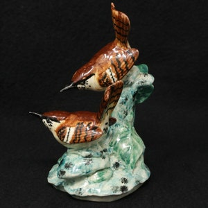 May include: Ceramic figurine of two brown and white birds perched on a green and blue leafy branch. The birds have detailed markings and are positioned facing different directions.