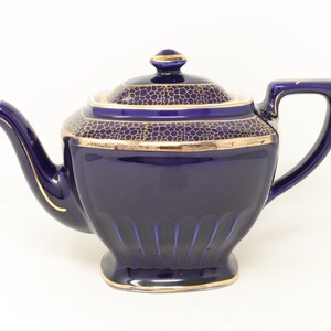 May include: A navy blue ceramic teapot with a gold trim and lid. The lid has a textured pattern. The teapot has a curved spout and handle. The teapot is approximately 7 inches tall.