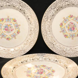 May include: Three vintage porcelain plates with a cream-colored background. The plates feature a gold filigree border and a central floral design in pink, blue, and yellow. The plates are arranged on a black surface.