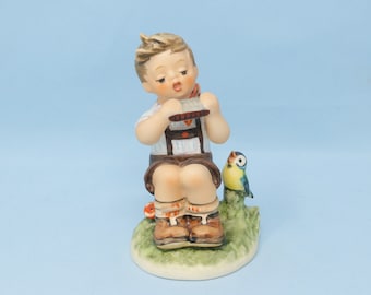 May include: A porcelain figurine of a boy playing a harmonica, sitting on a green base with a small blue bird perched on a branch. The boy is wearing a white shirt with a brown vest and brown shorts.