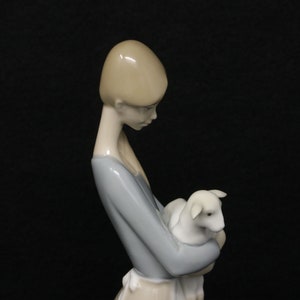 May include: A porcelain figurine of a young woman holding a lamb. The woman wears a light blue dress with a white apron. The lamb is white. The figurine has a smooth, glossy finish and is set against a black background.