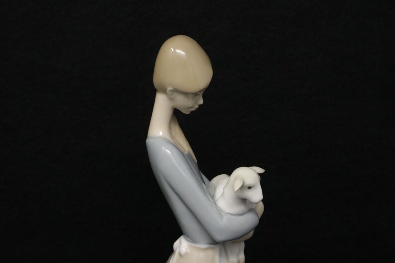 May include: A porcelain figurine of a young woman holding a lamb. The woman wears a light blue dress with a white apron. The lamb is white. The figurine has a smooth, glossy finish and is set against a black background.