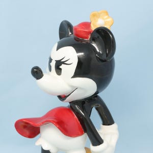 Puede incluir: Figura de Minnie Mouse de cer&aacute;mica, con cabeza y orejas negras, cara blanca y lazo rojo. Viste un vestido rojo y una flor amarilla en la cabeza. La figura est&aacute; sobre un fondo azul claro.