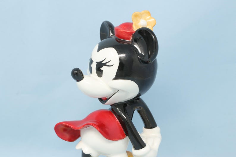 Puede incluir: Figura de Minnie Mouse de cer&aacute;mica, con cabeza y orejas negras, cara blanca y lazo rojo. Viste un vestido rojo y una flor amarilla en la cabeza. La figura est&aacute; sobre un fondo azul claro.
