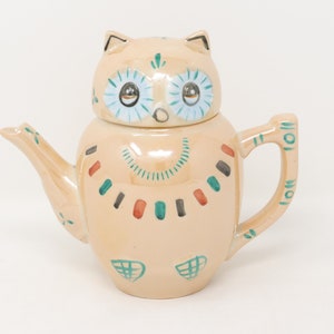 May include: A ceramic teapot shaped like an owl with a light brown body and green, blue, and red painted details. The owl has large blue eyes with silver pupils.