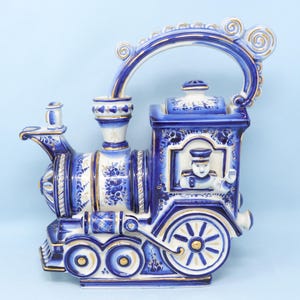 May include: A blue and white ceramic teapot shaped like a train. The teapot features gold accents and a detailed design, including a conductor figure in the window. The handle is arched and decorated with swirls.