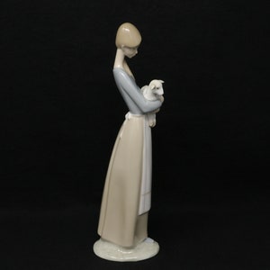 May include: A porcelain figurine of a woman holding a lamb. The woman wears a long, light-colored dress with a blue top and a white apron. The lamb is white. The figurine is on a white base.