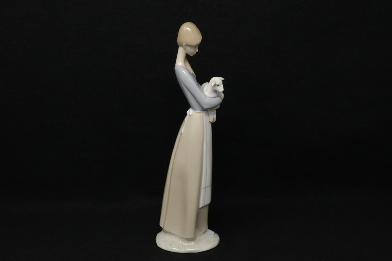 May include: A porcelain figurine of a woman holding a lamb. The woman wears a long, light-colored dress with a blue top and a white apron. The lamb is white. The figurine is on a white base.