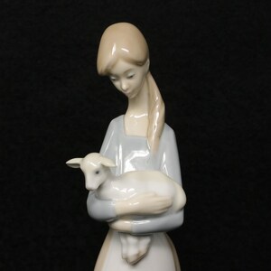 May include: A porcelain figurine of a woman holding a lamb. The woman wears a light blue dress and has long, light-colored hair. The lamb is white and rests in her arms. The figurine has a smooth, glossy finish and is set against a black background.