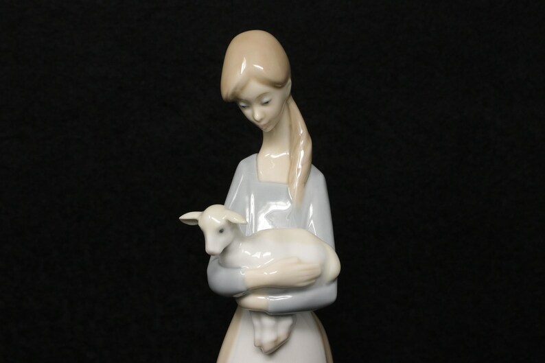 May include: A porcelain figurine of a woman holding a lamb. The woman wears a light blue dress and has long, light-colored hair. The lamb is white and rests in her arms. The figurine has a smooth, glossy finish and is set against a black background.