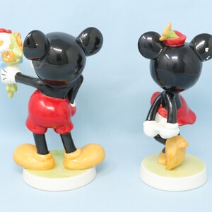Puede incluir: Dos figuras de cer&aacute;mica de Mickey y Minnie Mouse. Mickey sostiene un ramo de flores, viste pantalones cortos rojos y zapatos amarillos. Minnie lleva un vestido rojo y zapatos amarillos. Ambos tienen cuerpos negros y bases blancas.