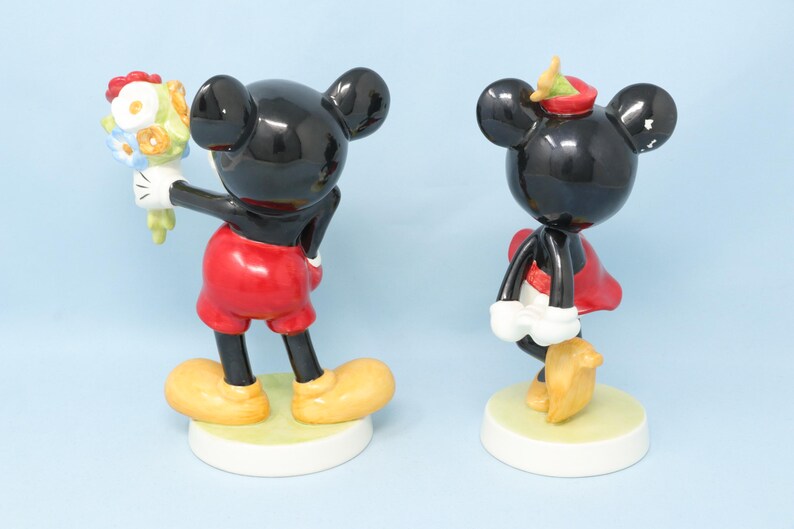 Puede incluir: Dos figuras de cer&aacute;mica de Mickey y Minnie Mouse. Mickey sostiene un ramo de flores, viste pantalones cortos rojos y zapatos amarillos. Minnie lleva un vestido rojo y zapatos amarillos. Ambos tienen cuerpos negros y bases blancas.