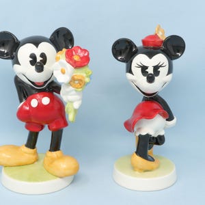 Puede incluir: Dos figuras de cer&aacute;mica de Mickey y Minnie Mouse. Mickey sostiene un ramo de flores, con pantalones cortos rojos y zapatos amarillos. Minnie lleva falda y sombrero rojos, con zapatos amarillos. Ambos tienen orejas negras y base blanca.