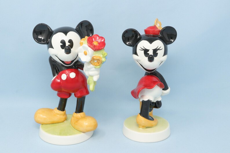 Puede incluir: Dos figuras de cer&aacute;mica de Mickey y Minnie Mouse. Mickey sostiene un ramo de flores, con pantalones cortos rojos y zapatos amarillos. Minnie lleva falda y sombrero rojos, con zapatos amarillos. Ambos tienen orejas negras y base blanca.