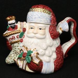 May include: A ceramic Santa Claus teapot with a red hat and beard. The teapot features a handle and spout, and is decorated with Christmas-themed details, including a stocking and gifts. The teapot is white, red, and gold.