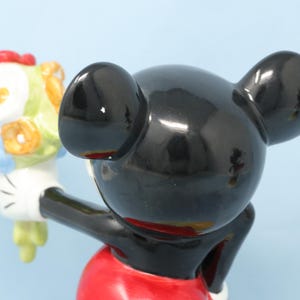 Puede incluir: Figura de cer&aacute;mica de Mickey Mouse sosteniendo un ramo de flores. La figura tiene una cabeza y orejas negras brillantes, un torso rojo y guantes blancos. El ramo presenta flores en tonos rojos, amarillos, azules y blancos.