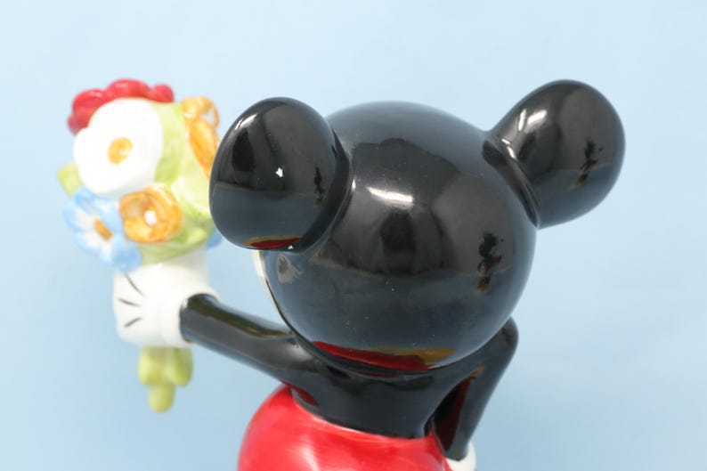 Puede incluir: Figura de cer&aacute;mica de Mickey Mouse sosteniendo un ramo de flores. La figura tiene una cabeza y orejas negras brillantes, un torso rojo y guantes blancos. El ramo presenta flores en tonos rojos, amarillos, azules y blancos.