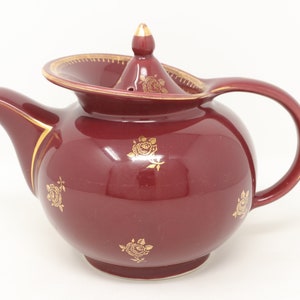 May include: A burgundy ceramic teapot with gold detailing on the lid, handle, and spout. The teapot features gold rose motifs. The lid finial is also gold. This teapot is designed for brewing tea.