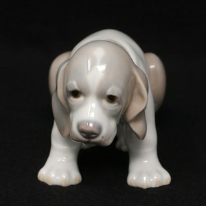 May include: A white porcelain figurine of a puppy with brown ears and brown spots on its face. The puppy is sitting with its head down and its paws tucked under its body.