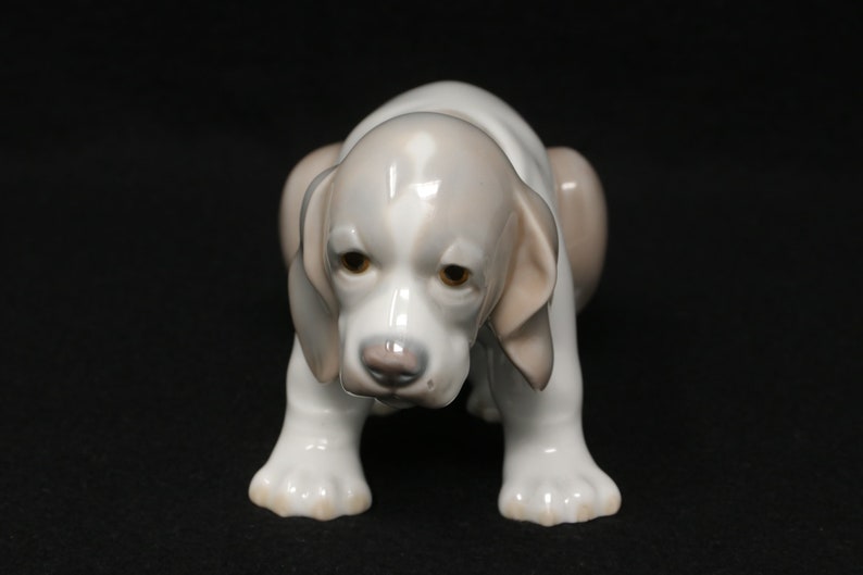 May include: A white porcelain figurine of a puppy with brown ears and brown spots on its face. The puppy is sitting with its head down and its paws tucked under its body.