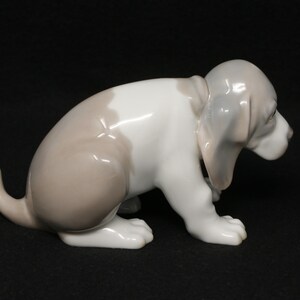 May include: A porcelain figurine of a white and brown puppy sitting with its head down. The puppy has floppy ears and a long tail.
