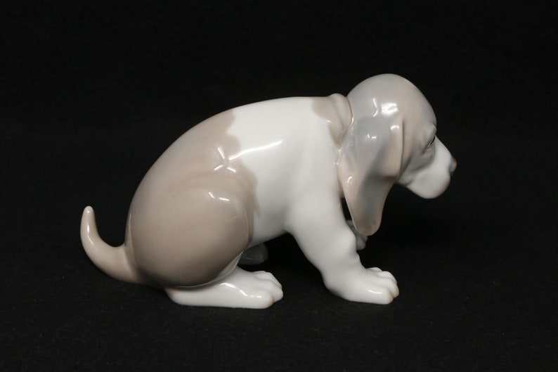 May include: A porcelain figurine of a white and brown puppy sitting with its head down. The puppy has floppy ears and a long tail.