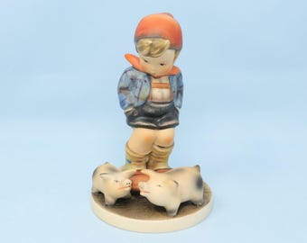 May include: A porcelain figurine of a young child in a blue jacket, orange hat, and black shorts, standing over two small pigs. The figurine is on a round, beige base, with a light blue background.