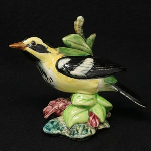 May include: A ceramic figurine of a yellow and black bird perched on a green leafy base with red flowers. The bird has a long tail and a black and white patterned wing.