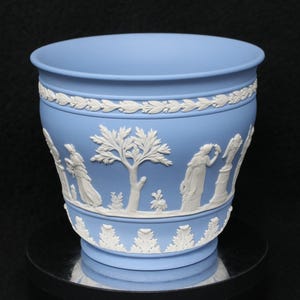 May include: A light blue ceramic planter featuring white relief decorations of classical figures, trees, and ornate leaf borders. The vessel has a flared rim and a tapered base, showcasing detailed molded scenes around the exterior surface.