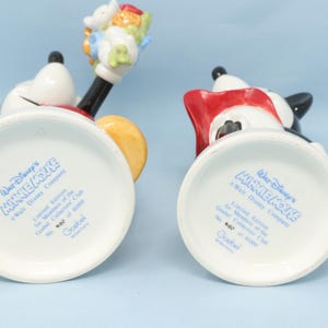 Puede incluir: Dos figuras de cer&aacute;mica de Minnie Mouse, una con flores. Las figuras son blancas, negras, rojas y amarillas. La parte inferior de cada figura tiene el texto "Walt Disney's MINNIE MOUSE".