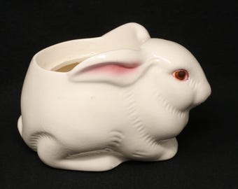 May include: A white ceramic bunny-shaped planter with a hollow interior. The bunny has pink-lined ears and reddish-brown eyes. The planter is designed to sit upright, with a textured surface and a rounded shape, perfect for small plants.