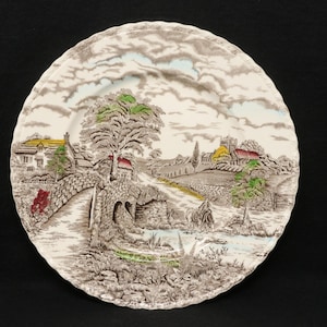 May include: A decorative ceramic plate with a landscape scene. The plate features a bridge, houses, trees, and a river, all in shades of brown, green, yellow, and blue. The rim of the plate has a scalloped edge.