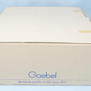 Puede incluir: Una caja de cart&oacute;n beige con la palabra azul "Goebel" y el texto "Bringing quality to life since 1871". La caja est&aacute; sellada con cinta adhesiva y tiene un fondo azul claro.