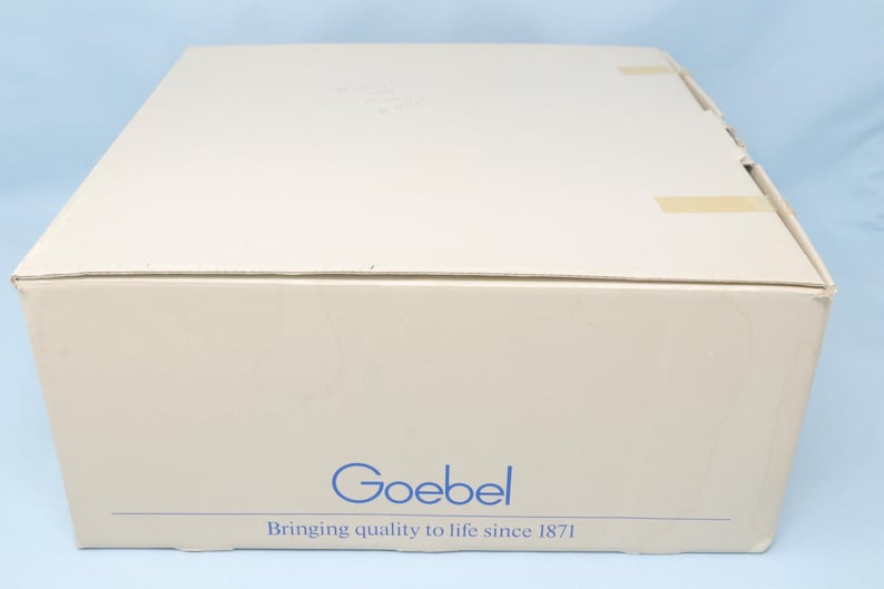 Puede incluir: Una caja de cart&oacute;n beige con la palabra azul "Goebel" y el texto "Bringing quality to life since 1871". La caja est&aacute; sellada con cinta adhesiva y tiene un fondo azul claro.