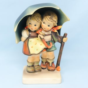 May include: A ceramic figurine of two children standing together under a large blue umbrella. The girl wears a yellow polka-dot apron over a brown dress, while the boy wears green shorts and a brown jacket. They stand on a rectangular base.