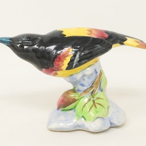 May include: A ceramic figurine of a black and yellow bird perched on a branch with green leaves. The bird has a red breast and a blue beak.