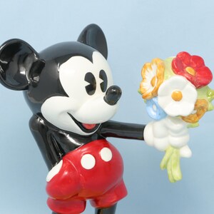 Puede incluir: Figura de cer&aacute;mica de Mickey Mouse sosteniendo un ramo de flores. Mickey est&aacute; representado en sus colores cl&aacute;sicos: negro, blanco, rojo y amarillo. El ramo incluye flores rojas, blancas, amarillas y azules.