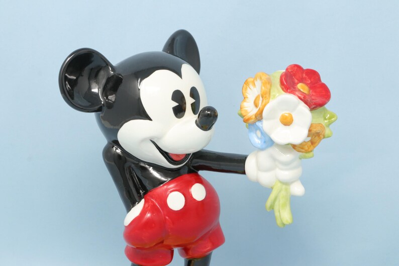 Puede incluir: Figura de cer&aacute;mica de Mickey Mouse sosteniendo un ramo de flores. Mickey est&aacute; representado en sus colores cl&aacute;sicos: negro, blanco, rojo y amarillo. El ramo incluye flores rojas, blancas, amarillas y azules.