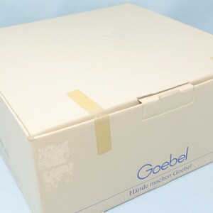 Puede incluir: Una caja de cart&oacute;n beige sellada con cinta adhesiva. La caja tiene la palabra "Goebel" impresa en azul, junto con la frase "H&auml;nde machen Goebel". La caja est&aacute; sobre una superficie azul claro.