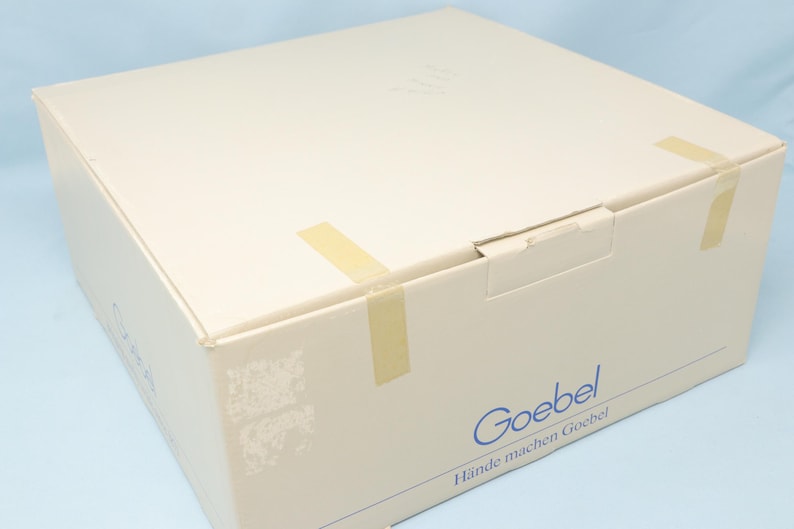 Puede incluir: Una caja de cart&oacute;n beige sellada con cinta adhesiva. La caja tiene la palabra "Goebel" impresa en azul, junto con la frase "H&auml;nde machen Goebel". La caja est&aacute; sobre una superficie azul claro.