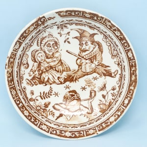 May include: A round, white ceramic plate with a brown transferware design. The plate features a central scene with figures and a border of floral and scroll patterns. The design includes a jester, a woman, a child, and a frog smoking a pipe.